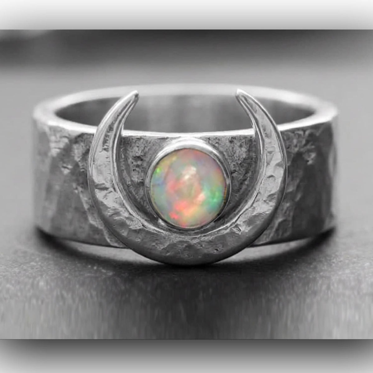 A gothic celestial silver ring handmade by SilverfireUK  featuring a textured moon crater band and a crescent moon design surrounding a round iridescent opal gemstone, shown on a dark textured surface.