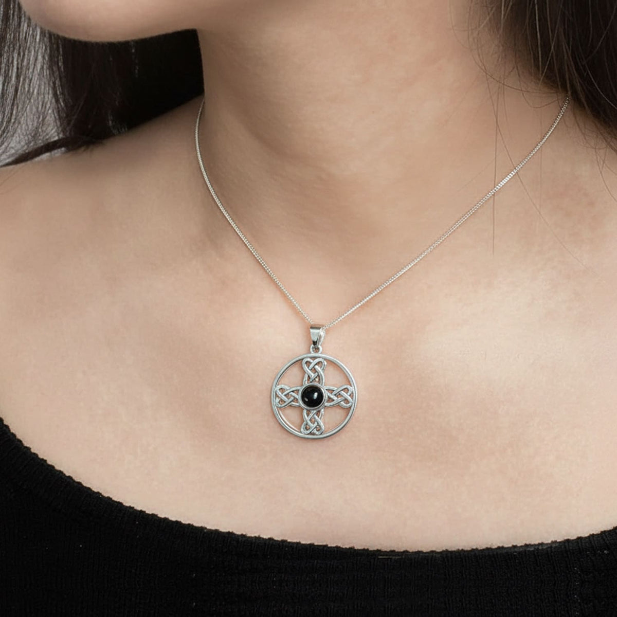 A close-up of a model wearing a silver Celtic knot cross pendant necklace with a central black onyx gemstone, paired with a black top.