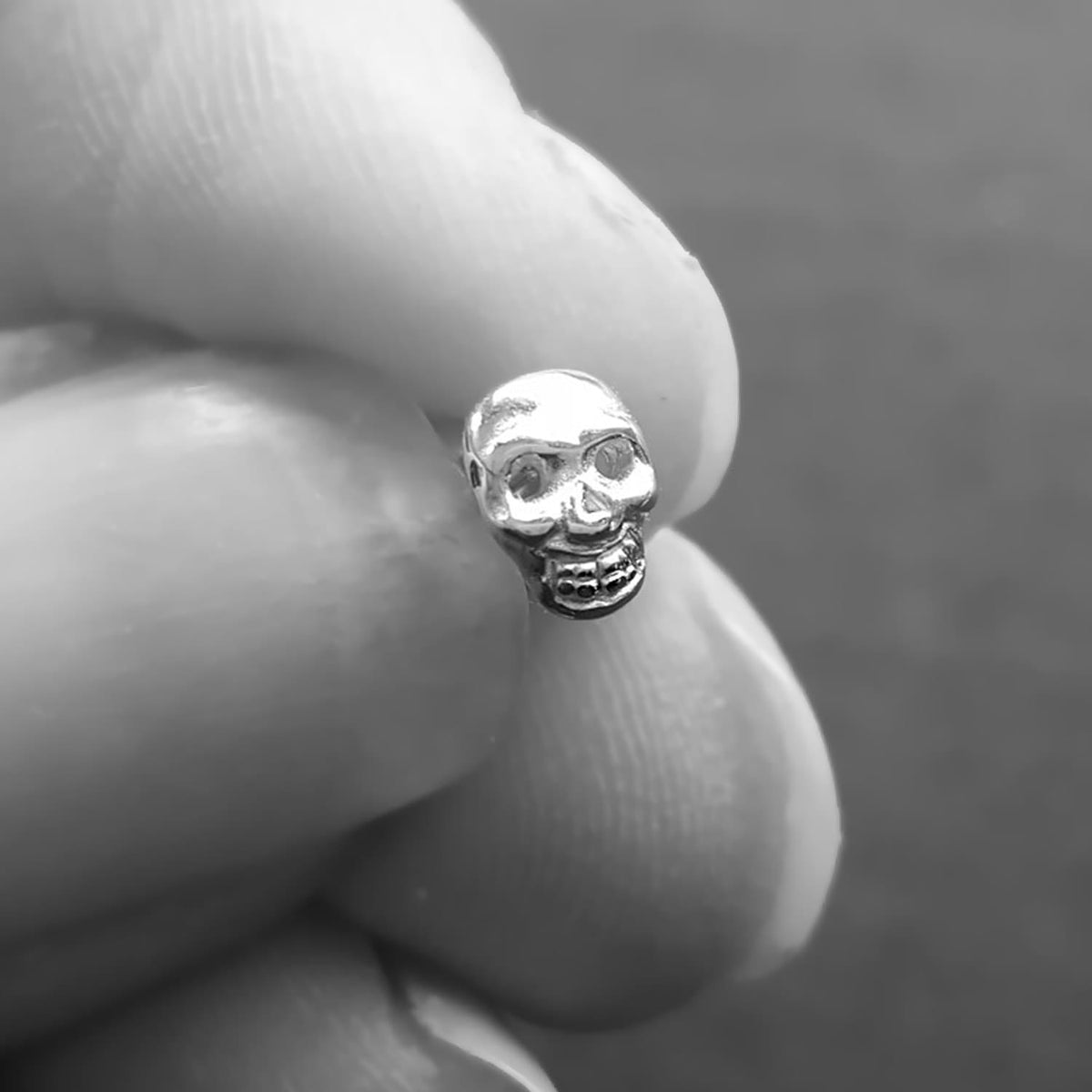 A small silver skull stud earring held between two fingers to show its scale, photographed in a close-up black and white style.